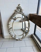 Full view of the vintage oval mirror leaning against wall and window, showing the entire frame and reflective surface.