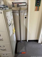 Full view of the medical scale showing the platform, vertical height rod, and beam balance mechanism positioned against a wall next to a filing cabinet.