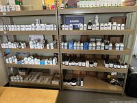 Two metal shelving units filled with supplement bottles and containers on four shelves each. Shelves show beige/tan metal structure, stocked with white, dark brown, and blue bottles mostly arranged in rows.
