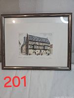 Framed signed drawing of European village street architecture hung on a wall, showing the timber-framed building with stone and wooden details