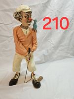 Front view of golfer sculpture wearing orange sweater, white cap, holding golf club with small bird on shoulder, white pants, and golf shoes.