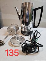 Full view of coffee percolator showing stainless steel body, black handle, detachable metal plunger, lid with glass knob, and attached power cord.