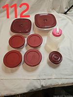 Top-down view of kitchen containers showing seven round glass bowls with red lids, two rectangular plastic containers with red lids, and a plastic squeeze bottle with a red cap.