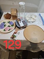 Lot of kitchen items on table including decorative floral plate, wooden lazy susan, glass pitcher, egg cooker, knife block, and egg tray