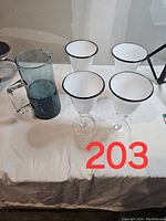 Photo showing four large white margarita glasses with black rims and a tall blue glass pitcher on a white surface.