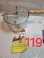 Closed view of the Whirley-Pop popcorn maker showing metal pot, hinged lid with wooden knobs, wooden handle, and included pamphlet.