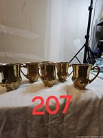 Front angle view of six gold-coloured espresso cups arranged in a semi-circle on a white surface with a wall and tripod in the background.