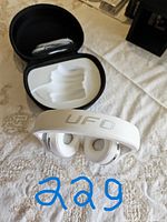 White Bluedio UFO wireless headphones displayed with cushioned ear cups and headband, laid on a white embroidered fabric surface. Headphones partially inside an open black carrying case with internal padding.