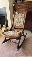 Full view of antique folding rocking chair showing wood frame, floral fabric seat and backrest, and carved floral detail on top backrest.