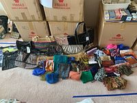 Photo showing multiple bags laid on carpet floor along with many folded scarves in different colors and prints, plus large cardboard boxes behind.