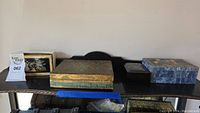 Set of four decorative jewelry boxes arranged on a black table. From left to right: small floral bird motif box, gold-tone textured box with floral inlay lid, blue stone-like patterned box on the right, and a larger black base box with mirrored interior below.
