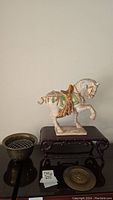 Side view of ceramic horse figurine placed on hardwood stand with brass flower pot and decorative brass disk nearby
