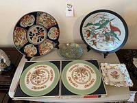Photo showing five decorative plates arranged on table including large warrior scene plate, round plate with floral and abstract designs, smaller floral plate, two boxed plates with floral borders, and two square floral plates with gold trim. Shows crazing and some chips.