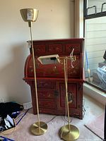 Two metal floor lamps shown side by side near a window and red Chinese lacquer desk. Taller lamp has a round base and a wide bowl-shaped shade. Shorter lamp has an adjustable arm with a rectangular shade and pull chain.