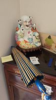 View of the Buddha piggy bank sitting on a wooden surface next to a folded blue and beige parasol and artwork.