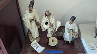 Group photo of three Shekwan porcelain figurines on a shelf with a man in white robes holding flowers, a seated man holding a scroll, and a man with a cracked bowl.