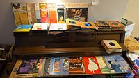 View of approximately 20 vinyl records stacked and spread on piano, covers facing forward showing various artists and genres including jazz and pop.