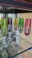Set of 5 colorful crystal wine glasses on glass shelf in varying colors (green, purple, amber, dark green, pink)