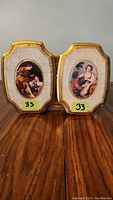 Two ornamental wood panels with oval prints, one showing a seated man and woman, the other a seated woman. Panels have gold edges and fabric-textured backgrounds.