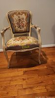 Front view of the antique chair showing embroidered floral fabric on backrest and seat, off-white painted wooden frame with minor wear.