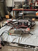 Battery charger and additional battery equipment with booster cables and cables laid out on white surface in garage or workshop area.