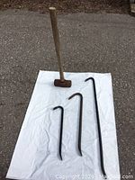 Photo of a 10 lb sledge hammer with wooden handle and two metal crowbars laid out on a white sheet on pavement.