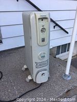 Full view of white NOMA electric oil-filled heater placed on a concrete surface outdoors. The heater stands upright with wheels visible.