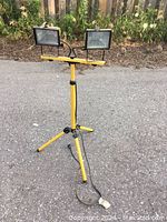 Dual flood lights mounted on an adjustable yellow tripod stand, placed on outdoor pavement with plants in background.