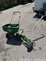 Side view of the green Scotts Turf Builder EdgeGuard DLX seed and salt spreader and the metal reel hand mower on asphalt.
