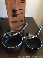 Image showing two black non-stick saucepans with glass lids and black handles, displayed on a table alongside the original box labeled 'KUEHN Nonstick Saucepan Set'.