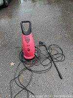 Photo of a red electric pressure washer with coiled black hose and spray wand resting on a wet asphalt surface.