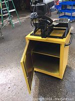 Yellow wheeled cabinet with open door showing internal storage shelves, base for mounting saw