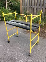Photo of a portable scaffold with yellow metal frame, black platform boards, and caster wheels on pavement outdoors.