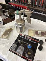 Metal spray gun with attached container alongside wrapped air brush kit and sanding discs on a white surface.