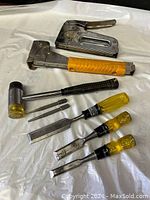 Photo showing two staplers, one chisel hammer, two nail punches, and three chisels with yellow handles laid out on a white surface.