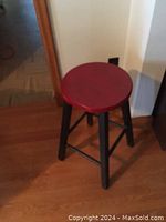 Photo of wooden stool with round red seat and black legs standing on a wooden floor near a white wall.