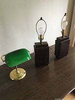 Photo of three lamps including two tall tri lamps with dark wooden bases and a green banker-style desk lamp on a wooden surface.