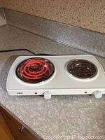 White two burner electric cooktop on countertop. Left burner is glowing red hot, right burner is off or warming.