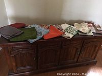 Photo showing a spread of assorted napkins and table cloths on a wooden cabinet, including solid color napkins and patterned table cloths.