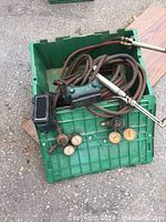 Green plastic crate containing multiple welding torches with hoses, pressure regulator gauges, and a black welding goggles mask on top.