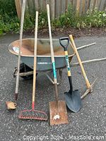 Metal wheelbarrow in used condition with wooden handles and single front wheel, surrounded by five garden tools.