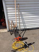 Wide shot of shovel, branch lopper, rake, electric chainsaw, and electric hedge trimmer on ground near garage door.