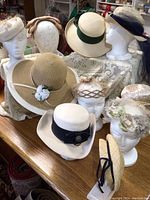 Photo showing 10 vintage hats on white mannequin heads and arranged on table, highlighting variety of styles and colors mainly in ecru and natural tones.