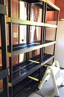 Two black Keter shelving units with five shelves each, grid-style shelving with yellow clips, standing next to windows with plaid curtains.