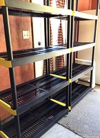 Two black plastic Keter shelving racks with yellow corner supports shown side by side indoors against wood paneling and near a window.