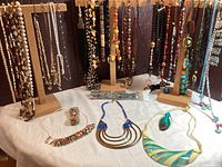 Overview of multiple necklaces including various bead and stone styles hanging on three wood stands, lying on white cloth backdrop