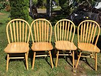 Four blonde oak chairs arranged side by side on grass, showing front view including spindle backrest, contoured seat, and legs with stretchers.