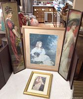 Four framed pictures placed on floor leaning against furniture: two tall narrow portraits of women, one larger portrait of a girl with dog, one smaller gold framed woman portrait