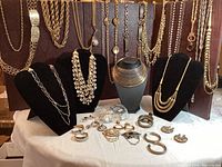 Overview photo showing various silver and gold-tone chain belts hanging on a display, multiple necklaces on stands, and earrings spread out on the table.
