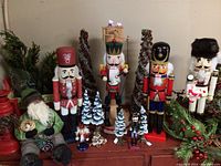 Photo showing large and small nutcracker figurines standing in a row with artificial snow-dusted pine trees, woodland Santa figurine seated, and red lanterns to the side.